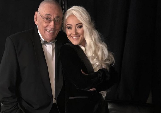 Maestrul Horia Moculescu zambitor,tinand mainile in buzunare, imbracat cu sacou negru si camasa alba, alaturi de fiica lui, Nidia, cu parul blond, imbracata in sacou negru de catifea si bluza alba