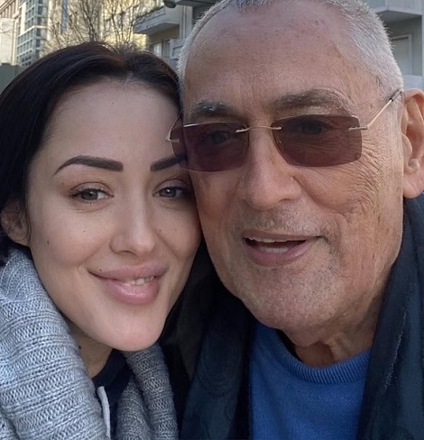 fotografie tip selfie, Nidia Moculescu zambitoare, imbracata in pulover gri, alaturi de tatal ei, Horia Moculescu, purtand ochelari si pulover albastru, in spatele lor aflandu-se cladiri