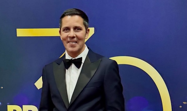 Lucian Viziru zambitor, imbracat cu sacou bleumarin, camasa alba si papion negru, in spatele lui aflandu-se un perete albastru cu cifre galbene pe care se vede cifra 30