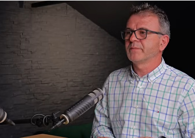 jurnalistul Mihai Stegerean imbracat cu camasa alba cu carouri verzi si violet, purtand ochelari si stand in fata microfonului in cadrul unui interviu 