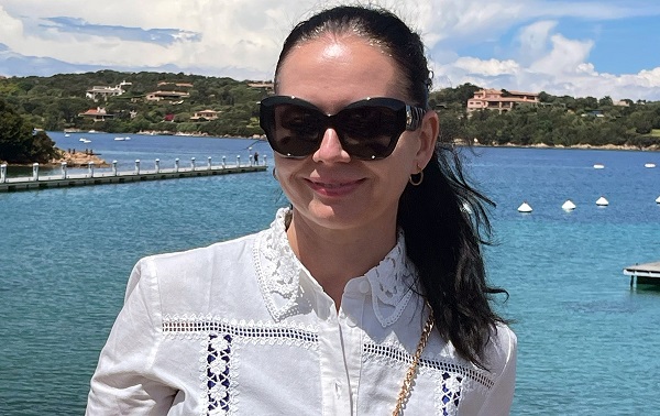 judecatoarea Ramona Mornaila zambitoare, imbracata cu bluza alba cu maneca lunga si broderie, purtand ochelari de soare negri, in timp ce se relaxa intr-o zona aflata la mare sau pe malul unui lac