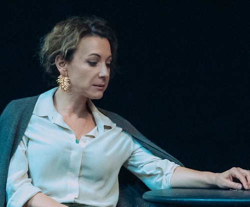 actrita Mirela Oprisor trista, pe fundal intunecat, imbracata in camasa alba cu maneca lunga si avand pe spate un pulover gri, purtand cercei aurii lungi si sprijinindu-si cotul de o masuta 