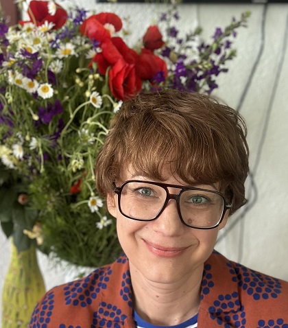 actrita Mirela Oprisor zambitoare, purtand ochelari, imbracata cu sacou maroniu cu imprimeuri albastre, avand in spatele ei un buchet cu maci si alte flori de c&acirc;mp 