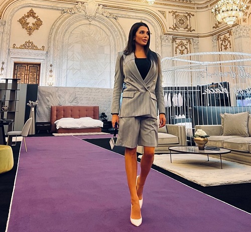 Elena Basescu imbracata intr-un costum gri,bluza neagra, cu sacou si pantaloni trei sferturi, purtand pantofi albi cu toc cui, in timp ce se afla la un show room cu produse de mobilier 