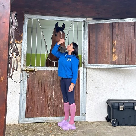 Elena Basescu imbracata in hanorac albastru, colanti negri, ciorapi roz si incaltaminte Crocks roz, in timp ce mangaie un cal aflat intr-un grajd la un centru de echitatie