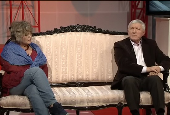 Diana Lupescu purtand bluza visinie, esarfa albastra, blugi si adidasi, Mircea Diaconu imbracat in costum maro inchis cu camasa alba, amandoi stand pe o canapea crem in timpul unei emisiuni de televiziune