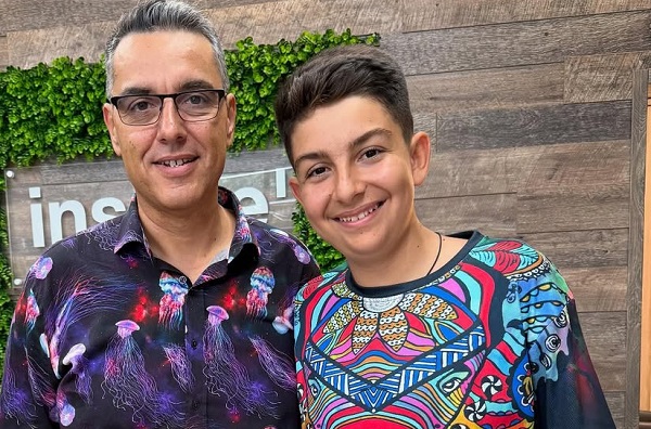 Cristi Brancu vesel, purtand ochelari, imbracat intr-o camasa cu imprimeu cu meduze colorate, alaturi de fiul lui, Tudor, imbracat intr-un tricou multicolor, stand amandoi pentru a fi fotografiati 