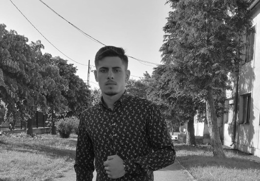 fotografie alb-negru cu Nelu Iulian Logofeteasa, &icirc;mbracat in camasa cu imprimeu cu maneca lunga, stand in preajma unui bloc langa care se afla copaci 