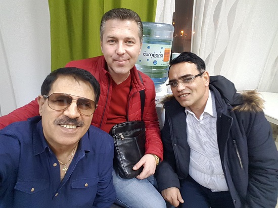 Dan Ciotoi vesel, purtand ochelari, imbracat cu camasa albastra descheiată la gat, avand la gat un medalion auriu cu cruce, stand alaturi de doi membri ai formatiei sale