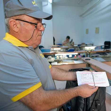 Postas in uniforma a Postei Romane, cu tricou bleu deschis si cu sapca inscriptionate cu galben cu simbolul Poștei, in timp ce are in mana cupoane de pensie scoase din geanta de postas in timp ce se afla la oficiul postal