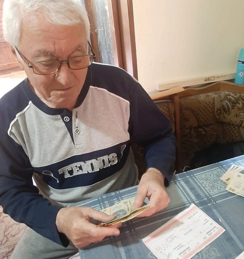 pensionar purtand ochelari de vedere, imbracat cu bluza de trening bleumarin cu gri, in timp ce sta la masa numarandu-si banii primiti la pensie si verificandu-si si cuponul de pensie
