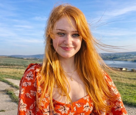 Andreea Anița, tanara de 27 de ani bolnava de cancer, zambitoare, imbracata intr-o rochie portocalie cu flori, in timp ce se afla in natura, in preajma unor dealuri si a unei ape