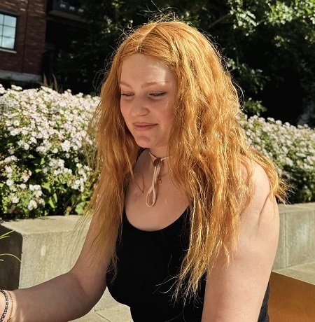 Andreea Anița, tanara de 27 de ani bolnava de cancer, imbracata cu top negru, privind in jos in timp ce se afla la o masuta in aer liber, in apropiere de tufisuri de flori si o cladire