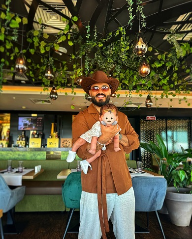 Jador imbracat cu camasa maro cu cordon in talie, pantaloni albi, purtand palarie maro pe cap si ochelari de soare, fericit in timp ce isi tine in brate baietelul, caruia nu ii dezvaluie chipul, langa barul dintr-un restaurant