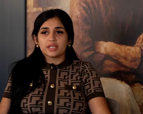 Melek Mincă, văduva victimei lui Mario Iorgulescu, imbracata cu bluza sau rochie maro cu imprimeu negru, cu nasturi aurii, purtand cercei aurii, in timp ce sta cu spatele la un tablou in timpul unui podcast la care a fost invitata