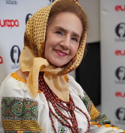 Sofia Vicoveanca zambitoare, imbracata in costum traditional, cu ie brodata cu verde, portocaliu, galben si maro, avand la gat un sirag de margele maronii, basma pe cap si stand pe scaun intr-un studio de televiziune sau la un post de radio