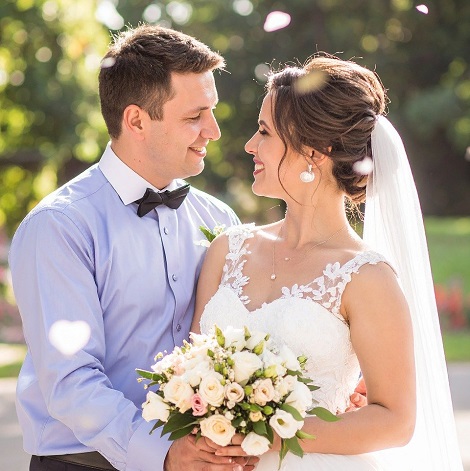 mire si mireasa intr-un spatiu in aer liber, mireasa in rochie alba cu umerii dezgoliti, cu val pe cap, tinand in mana un buchet de flori, uitandu-se la mire, imbracat cu camasa bleu cu guler alb si cu papion negru, zambind unul la altul