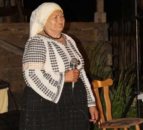 interpreta de muzica populara din Mehedinti Domnica Trop, imbracata in costum popular, cu ie brodata cu negru, fusta neagra si basma alba pe cap, tinand in mana un microfon in timp ce se pregateste sa cante seara, in aer liber, langa o casa taraneasca traditionala din lemn