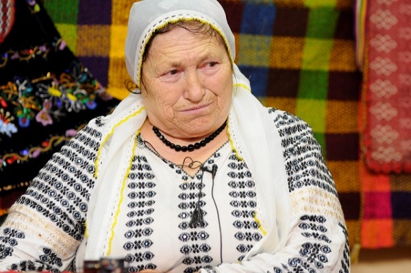 interpreta de muzica populara din Mehedinti Domnica Trop imbracata cu ie brodata cu negru, purtand margele negre la gat, basma alba cu broderie galbena pe cap, in spatele ei aflandu-se o haina neagra cu broderie colorata si paturi colorate