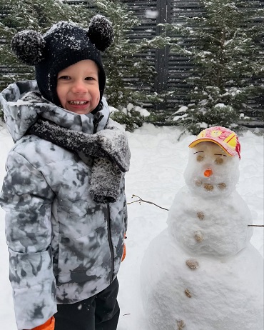 Tiago Luca, fiul lui Dani Oțil și al Gabrielei Prisacariu, imbracat cu geaca alb cu gri, cu pantaloni negri de f&acirc;ș, purtand pe cap caciula neagra cu urechi de ursulet, fular la gat, in timp ce se afla langa un om de zapada intr-o curte cu brazi