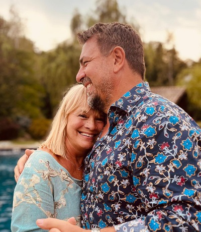 Horia Brenciu imbracat cu camasa inflorata pe fond inchis la culoare, fericit in timp ce isi imbratiseaza sotia, Alice Dumitrescu, imbracata cu rochie bleu cu flori albe, cei doi aflandu-se intr-un colt de natura, pe marginea unei ape 