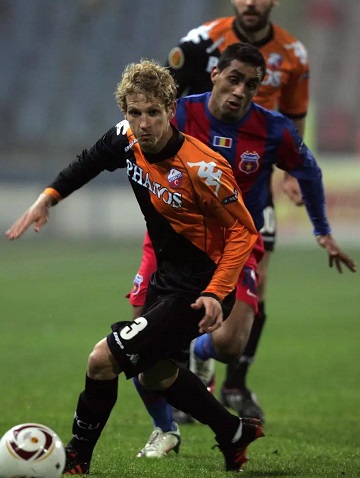 Mihai Neșu in prim-plan, pe terenul de fotbal, cu mingea in timpul unui meci, in timp ce in spatele lui se afla alti doi jucatori