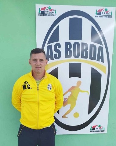 antrenorul Ciprian Manolache de la echipa de fotbal AS Bobda, imbracat cu bluza de trening galbena si cu pantaloni inchisi la culoare, stand rezemat de un perete pe care se afla insemnele clubului de fotbal si al firmei care il sponsorizeaza