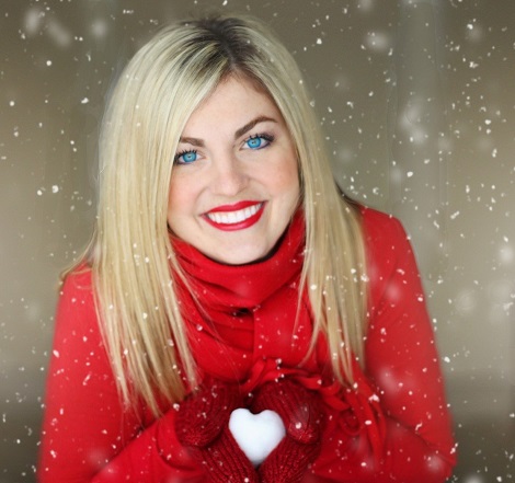 femeie frumoasa, blonda, imbracata in rosu, cu fular rosu si manusi rosii, zambind in timp ce tine in maini o inima de zapada intr-un cadru cu ninsoare