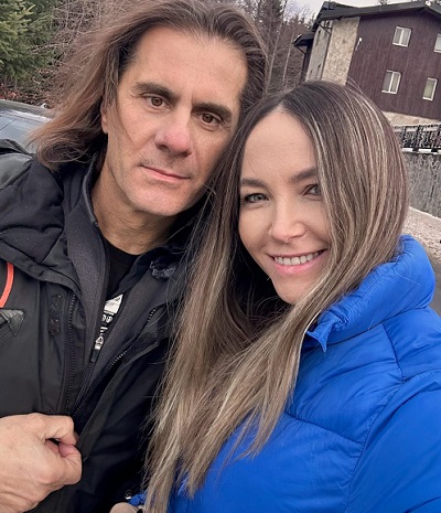Octavia Geamana imbracata cu geaca albastra, zambitoare, stand la pieptul sotului ei, Marian Ionescu, imbracat cu geaca neagra, amandoi aflandu-se langa o vila sau un hotel de la munte