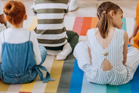 fetita cu parul roscat, cu coc, imbracata cu salopeta de blugi si bluza alba, stand pe un covoras cu dungi colorate langa alta fetita, cu codite, imbracata cu salopeta alba cu dungi bleu, in fata lor aflandu-se un alt copil cu tricou cu dungi si cu pantaloni oliv