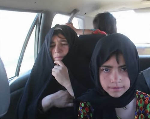 femeie afgana cu val negru, imbracata in negru, langa ea stand o fetita imbracata cu bluza colorata si cu val negru, iar in spatele lor fiind o fetita care se uita pe geamul masinii in care se afla toate trei