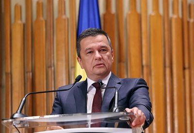 Sorin Grindeanu imbracat cu sacou bleumarin, camasa alba si cravata visinie, avand in spatele sau un steag albastru si stand la pupitru in fata microfoanelor in timpul unei interventii 