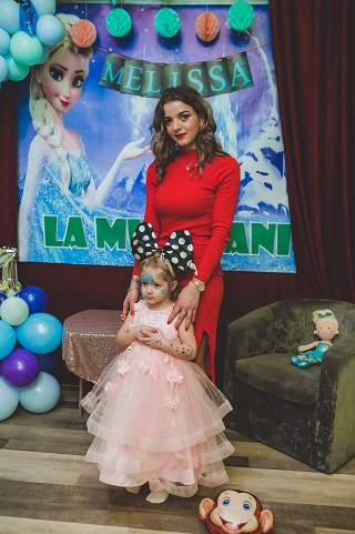 Corina Matras, tanara de 26 de ani decedata alaturi de fiica ei intr-un accident, imbracata cu rochie rosie si tinandu-si mainile pe umerii fetitei sale imbracata cu rochita roz cu volane de tul si purtand pe cap urechi de Minnie Mouse cu buline, cu ocazia aniversarii micutei