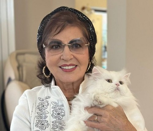 Irina Loghin imbracata cu bluza alba tip hanorac cu model floral negru pe piept, purtand bentita neagra lucioasa pe cap, cercei rotunzi aurii si ochelari, in timp ce isi tine in brate pisica alba acasa 