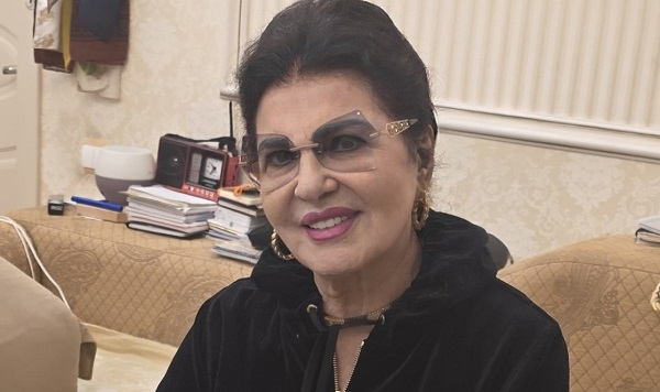 Irina Loghin imbracata cu hanorac de trening negru,cu cercei aurii si cu ochelari, stand pe o canapea crem si avand in spatele ei o masuta pe care se afla diverse obiecte si un perete bej deschis