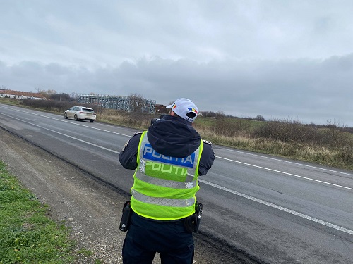 polițist din cadrul Poliției Rutiere Caraș-Severin, imbracat in uniforma, cu sapca alba pe cap, stand pe marginea unei sosele si incercand sa foloseasca un dispozitiv cu radar pentru a masura viteza masinilor care trec