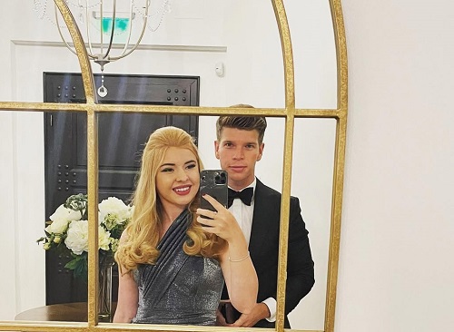 Karina Pavel imbracata cu rochie argintie, facandu-si selfie intr-o camera de hotel alaturi de partenerul ei, Dragos Iamandoiu, imbracata cu costum negru, camasa alba si papion