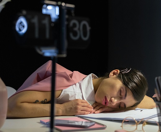 femeie tanara imbracata cu bluza alba fara maneci, avand pe spate un sacou roz, in timp ce doarme cu capul pe biroul plin de diverse rechizite la o ora tarzie la locul de munca