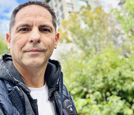 Dan Negru imbracat cu tricou alb si jacheta bleumarin, stand pe un spatiu verde in aer liber, in spatele sau fiind un copac si o cladire cu multe etaje