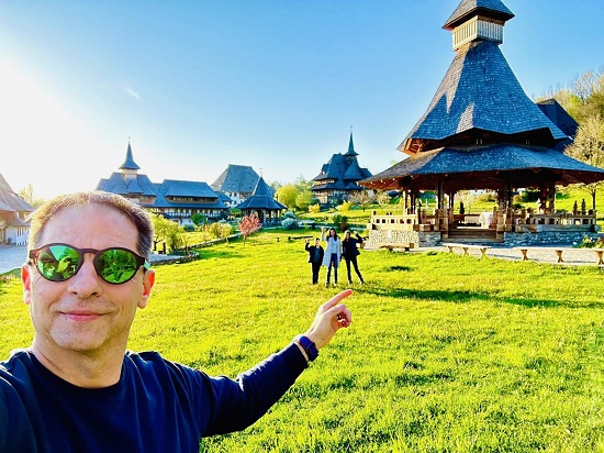 Dan Negru cu ochelari de soare, imbracat cu tricou bleumarin cu maneca lunga, aflandu-se pe o pajiste intr-un cadru cu biserici maramuresene, aratand cu degetul in spatele sau, unde se afla cei doi copii si sotia lui