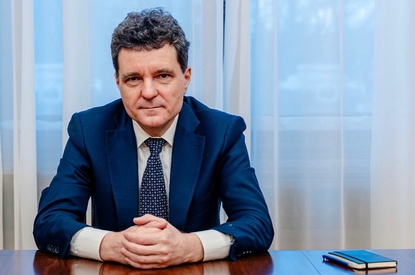 Președintele Rom&acirc;niei, Nicusor Dan, imbracat cu sacou bleumarin, camasa alba si cravata albastra stand pe scaun, cu palmele impreunate, asezate pe un birou, langa el aflandu-se un carnetel 