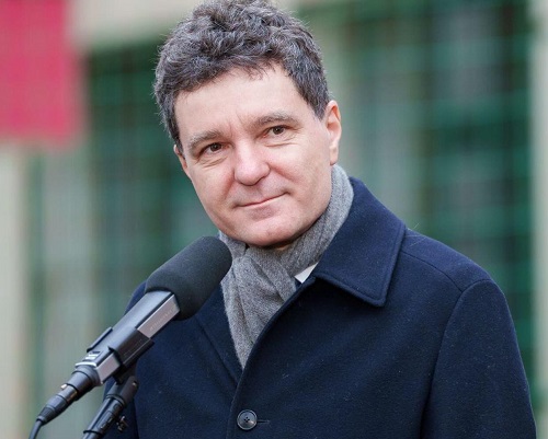 președintele Rom&acirc;niei Nicușor Dan zambitor, imbracat cu palton de stofa bleumarin, purtand fular gri, in timp ce vorbeste la microfon 