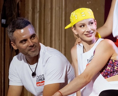 Sandra Izbasa veseala, imbracata cu un tricou alb fara maneci, purtand o sapca galbena, langa ea aflandu-se sotul ei, Razvan Banica, zambitor, pregatindu-se pentru competitia Power Couple