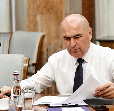 premierul Ilie Bolojan imbracat in camasa alba cu maneca lunga, cu cravata neagra, stand pe scaun și analizand diverse documente cu pixul in mana in timpul unei sedinte de lucru de la Guvern