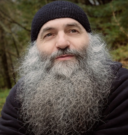 parintele Pimen Vlad ganditor, imbracat in hanorac negru, petrecandu-si timpul intr-un colt de natura cu copaci inverziti