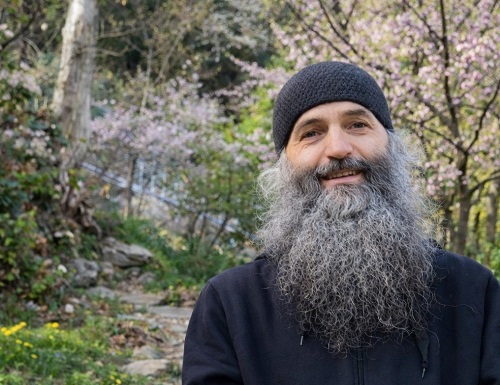 părintele Pimen Vlad zambitor, imbracat in hanorac negru, stand intr-un colt de natura cu copaci infloriti primavara