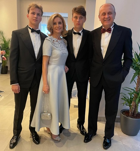 Diana Șucu imbracata in rochie eleganta lunga, gri perlat, cu sandale argintii si cu poseta argintie, fericita alaturi de sotul ei, Dan Șucu și de cei doi baieti ai lor, toti imbracati in costume negre, camasi albe si papion intr-o locatie de lux