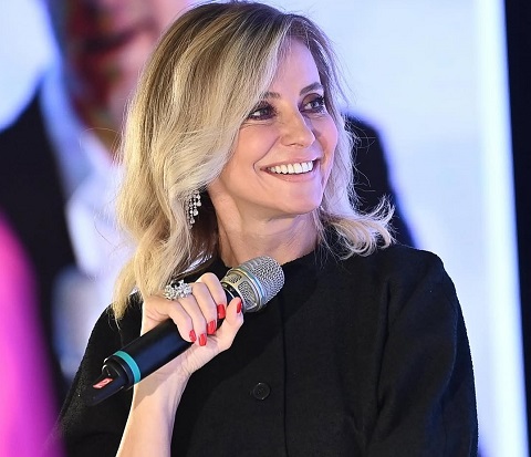 Diana Șucu fotografiata din profil, zambitoare,imbracata in negru, purtand cercei lungi din strasuri si un inel masiv cu argintiu si piatra neagra, in timp ce tine in mana un microfon