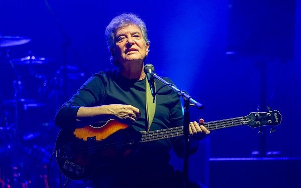 basistul trupei Holograf, Iulian Mugurel Vrabete,imbracat in negru, stand pe un scaun si cantand la chitara in fata microfonului pe scena in timpul unui spectacol 