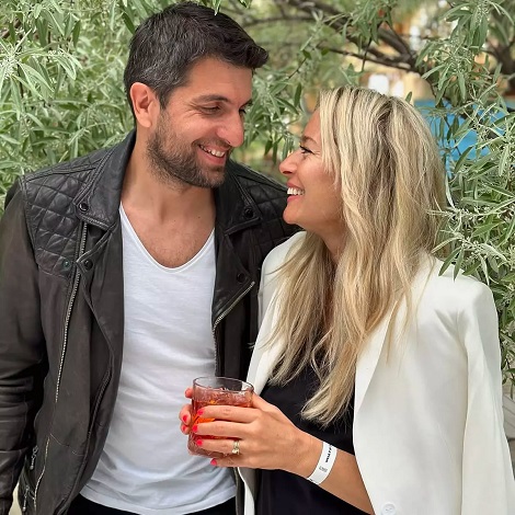 Laura Cosoi imbracata cu sacou alb, tinand in mana un pahar cu o bautura si uitandu-se zambind spre soțul ei, Cosmin Curticapean, imbracat cu tricou alb si geaca neagra de piele, amandoi aflandu-se in aer liber, langa un arbore